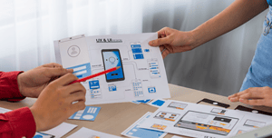 Due designer collaborano su documenti che rappresentano il layout di un'app mobile, enfatizzando l'importanza di menu di navigazione intuitivi nel design UX/UI.