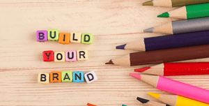 Parole colorate 'BUILD YOUR BRAND' con cubi lettera in primo piano e matite colorate sparse su uno sfondo in legno, illustrando la costruzione di un personal branding nell'ambito del web design.