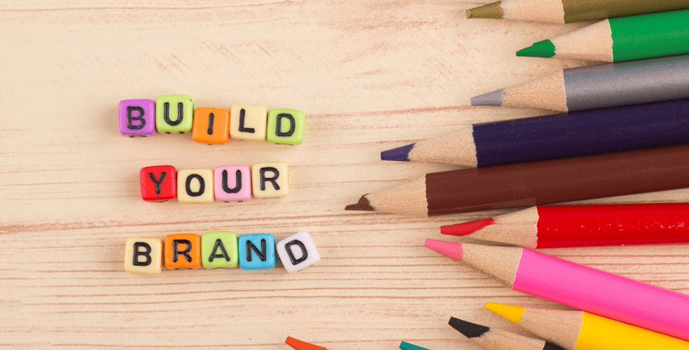 Parole colorate 'BUILD YOUR BRAND' con cubi lettera in primo piano e matite colorate sparse su uno sfondo in legno, illustrando la costruzione di un personal branding nell'ambito del web design.