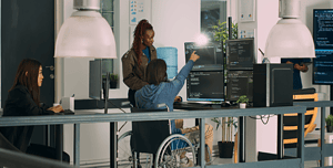 Una squadra di web designer, inclusa una persona in sedia a rotelle, lavora insieme davanti a monitor con codice di programmazione, enfatizzando l'accessibilità e l'inclusività nel web design.