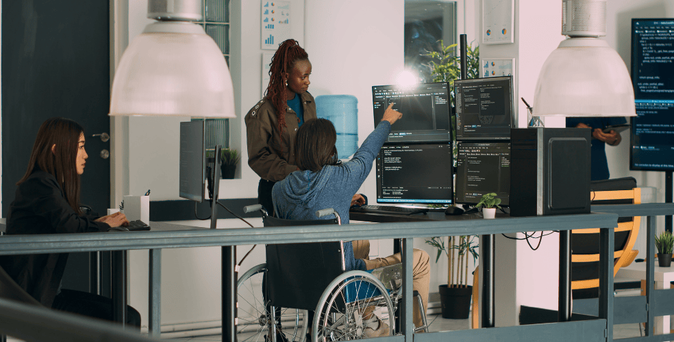 Una squadra di web designer, inclusa una persona in sedia a rotelle, lavora insieme davanti a monitor con codice di programmazione, enfatizzando l'accessibilità e l'inclusività nel web design.
