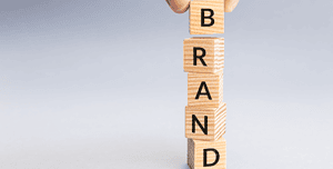 Una mano posiziona con precisione blocchi di legno incisi con le lettere che compongono la parola 'BRAND' su uno sfondo neutro, rappresentando la costruzione e la consistenza necessaria nel personal branding.