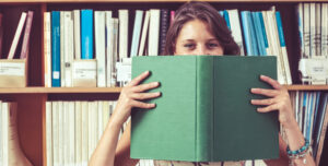 Donna che legge un grande libro verde, con una libreria piena di libri sullo sfondo.