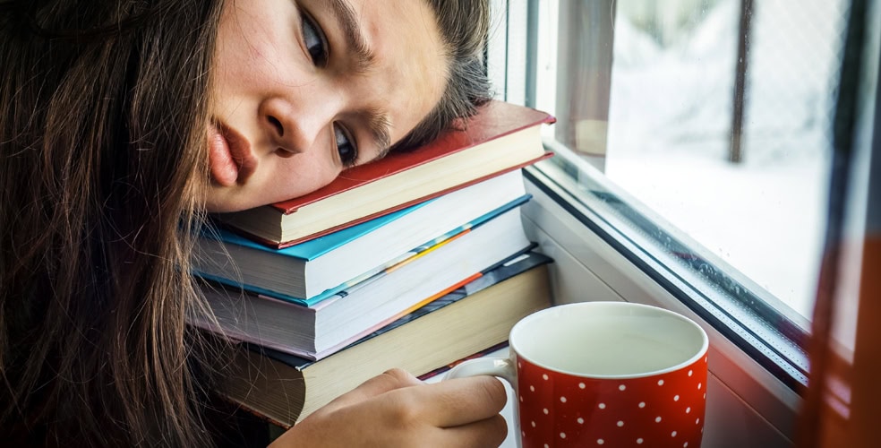 Giovane donna appoggiata su una pila di libri con un'espressione stanca e una tazza di tè accanto, guardando fuori dalla finestra, rappresentando le difficoltà di concentrazione durante la lettura.