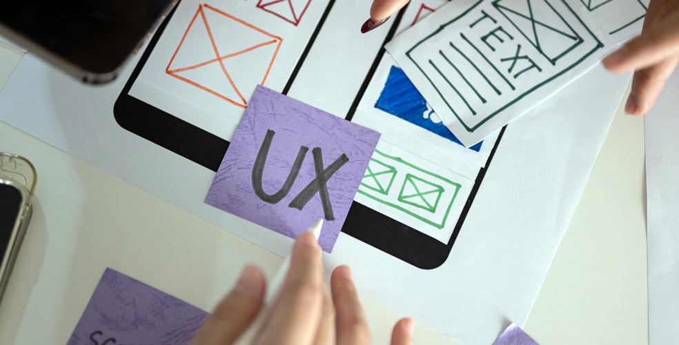 Mani che dispongono cartoncini con design UX e wireframe su un piano di lavoro, evidenziando il processo di sviluppo di un'interfaccia utente responsive.