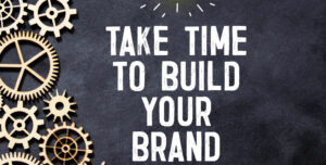 Ingranaggi di legno su uno sfondo scuro con la scritta 'Take time to build your brand', enfatizzando l'importanza del tempo e dell'attenzione per creare un marchio coerente.