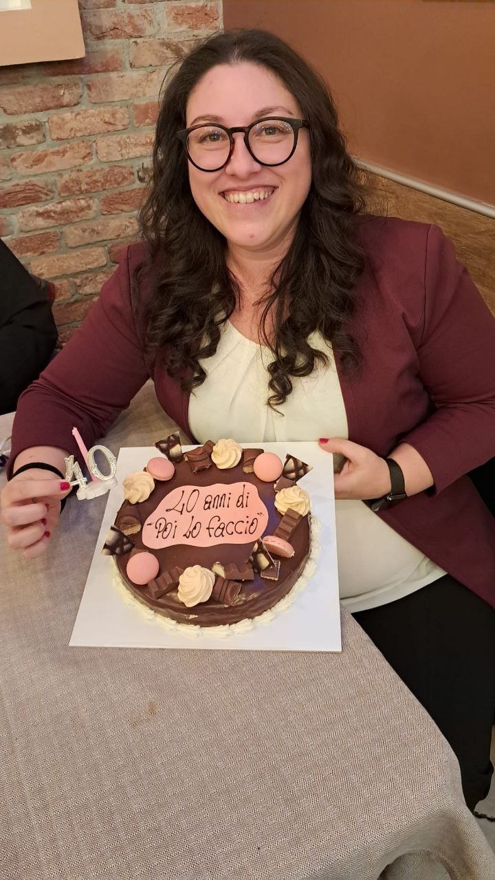 Una donna sorridente con capelli lunghi e occhiali celebra il suo compleanno, tenendo una torta al cioccolato decorata con macaron e cioccolatini. Sopra la torta è scritto "40 anni di 'poi lo faccio'". Sul tavolo, una candela rosa a forma di numero 40. L'immagine trasmette un momento di festa e riflessione sui traguardi raggiunti.