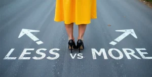 Donna in piedi al centro di una strada asfaltata con indosso un abito giallo e scarpe con tacco nere, davanti a due frecce opposte con le scritte “LESS” e “MORE”, che simboleggiano una scelta tra quantità e qualità, a rappresentazione visiva del concetto di ottimizzare il lavoro puntando sull'efficienza piuttosto che sull'aumento del carico, in linea con l’obiettivo di accogliere 13 nuovi clienti ideali nel 2025.