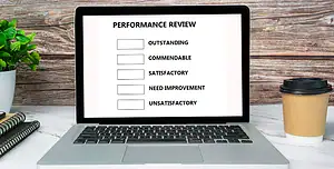 Laptop aperto su scrivania con modulo “PERFORMANCE REVIEW” a schermo: elenco di valutazioni con caselle da spuntare — Outstanding, Commendable, Satisfactory, Need Improvement, Unsatisfactory; accanto taccuini a righe, pianta verde e bicchiere da asporto per caffè; sfondo in legno rustico. Concetto di valutazione delle prestazioni, feedback HR e revisione periodica degli obiettivi in ufficio o in smart working.