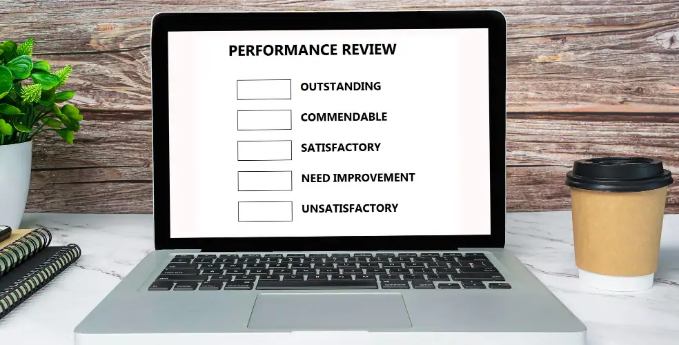 Laptop aperto su scrivania con modulo “PERFORMANCE REVIEW” a schermo: elenco di valutazioni con caselle da spuntare — Outstanding, Commendable, Satisfactory, Need Improvement, Unsatisfactory; accanto taccuini a righe, pianta verde e bicchiere da asporto per caffè; sfondo in legno rustico. Concetto di valutazione delle prestazioni, feedback HR e revisione periodica degli obiettivi in ufficio o in smart working.