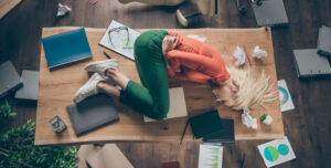Donna distesa in posizione fetale sopra una scrivania in disordine, circondata da documenti, cartelle e grafici sparsi, simbolo visivo di sovraccarico cognitivo e burnout legato alla mancanza di concentrazione e gestione efficace del lavoro profondo.