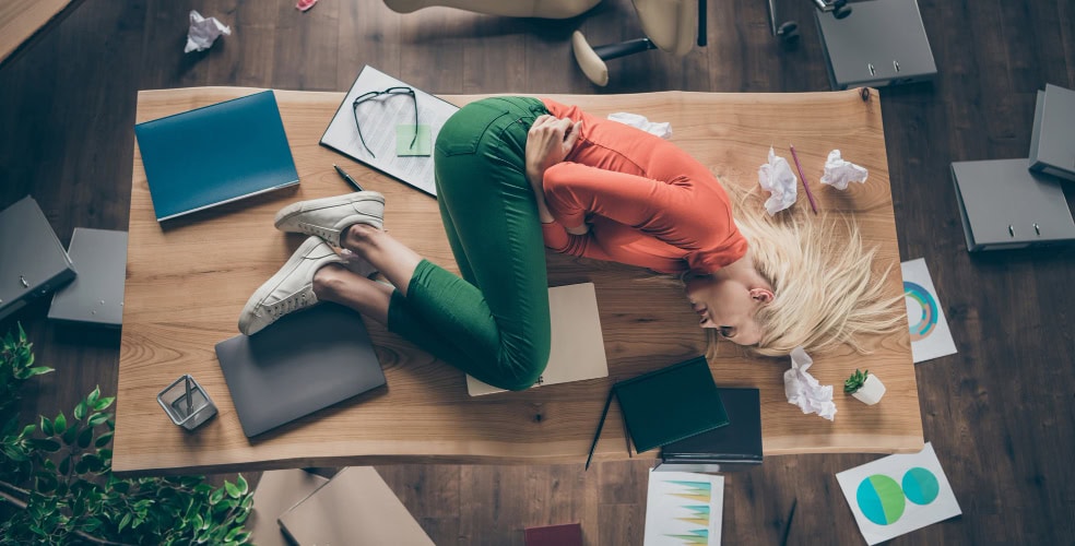 Donna distesa in posizione fetale sopra una scrivania in disordine, circondata da documenti, cartelle e grafici sparsi, simbolo visivo di sovraccarico cognitivo e burnout legato alla mancanza di concentrazione e gestione efficace del lavoro profondo.